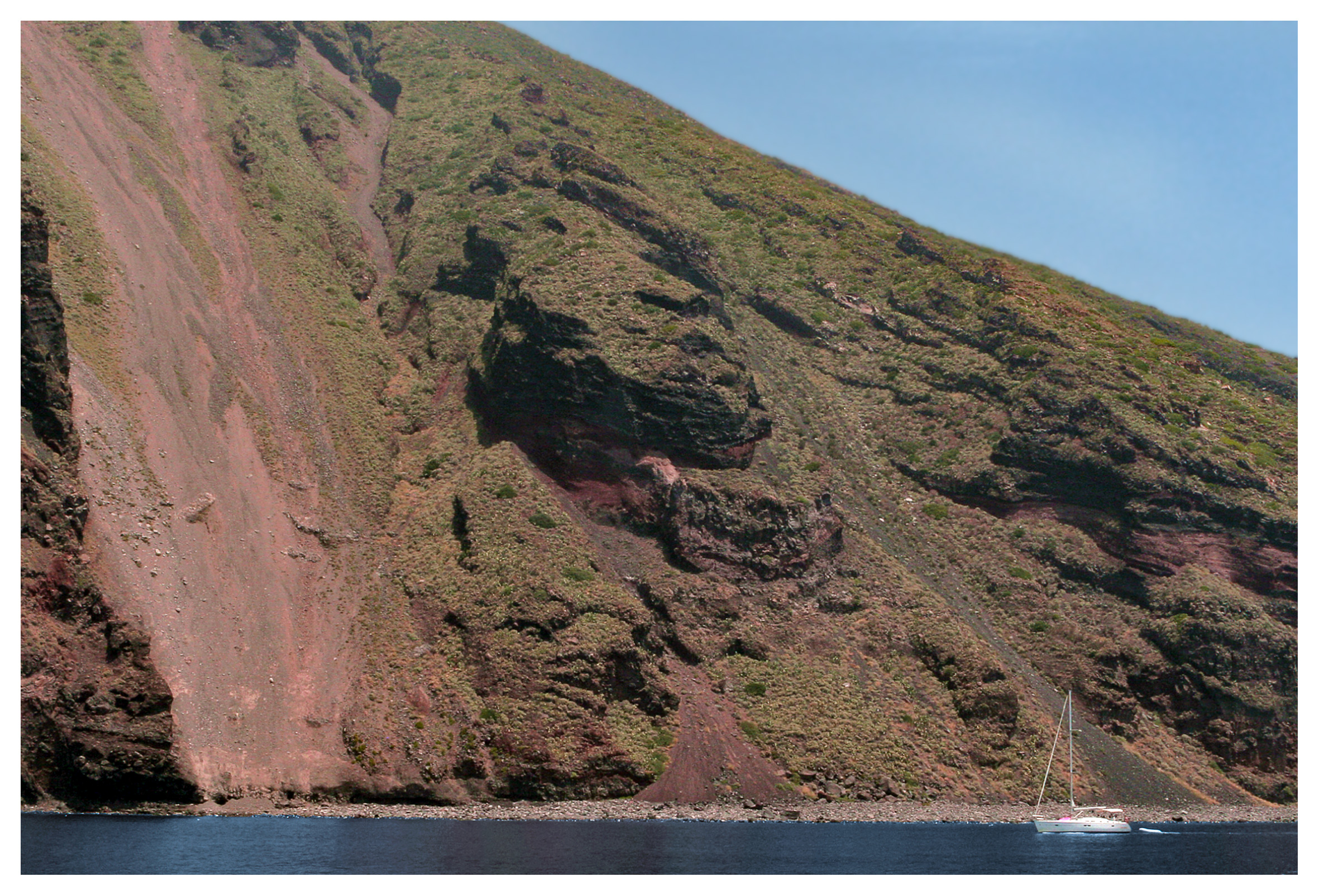 Yachting photography: A yacht sailing close to a towering volcanic hillside at Filicoudi, Aeolian Islands, Sicily